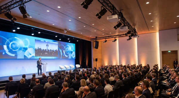Salle de conférence ou d'assemblée générale professionnelle moderne, avec une grande audience assise face à une scène. Au centre : un orateur à un pupitre, écran LED géant haute définition en arrière-plan affichant des contenus (graphiques, vidéo, présentation). Projecteurs de scène professionnels, vidéoprojecteurs visibles en hauteur. Système de visioconférence intégré avec caméra motorisée pointée sur l'orateur. Public captif, configuration de type assemblée ou séminaire corporate. Photographie réaliste, éclairage professionnel chaud, plan large capturant la configuration technique et l'interaction humaine.